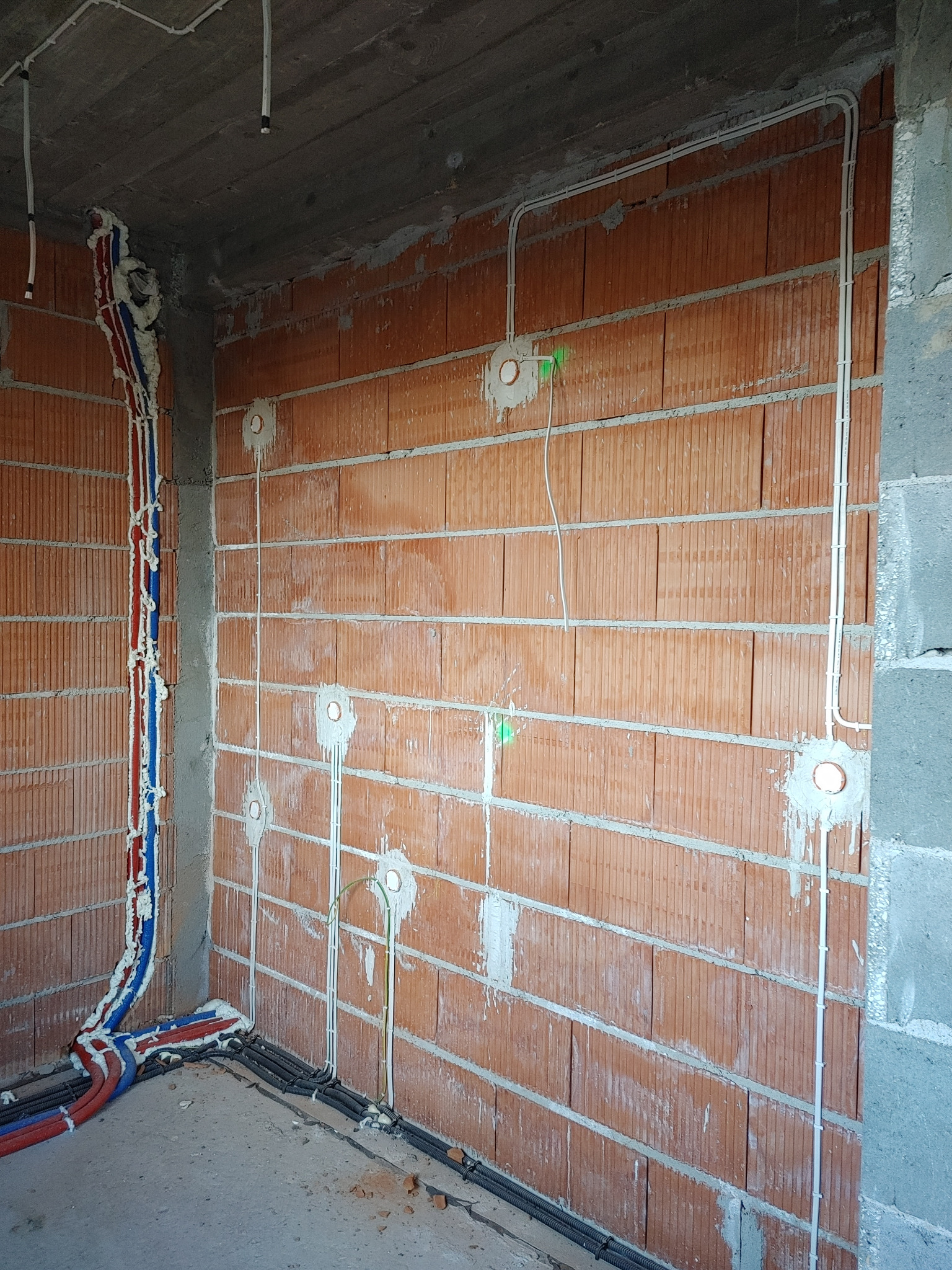 Wnętrze w trakcie prac instalacyjnych: widoczne przewody elektryczne w peszlach i rurkach mocowanych na ścianie z czerwonej cegły oraz rury instalacji wodnej zabezpieczone pianką montażową.