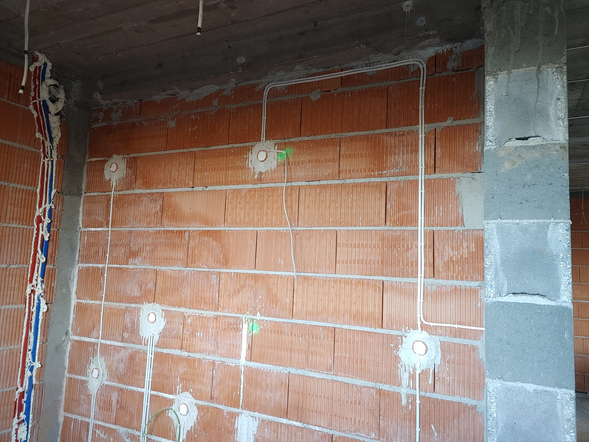 Surowy mur z cegły z widocznymi przewodami elektrycznymi w białych rurkach, osadzonymi gniazdkami i pionowymi rurami hydraulicznymi zabezpieczonymi pianką montażową.