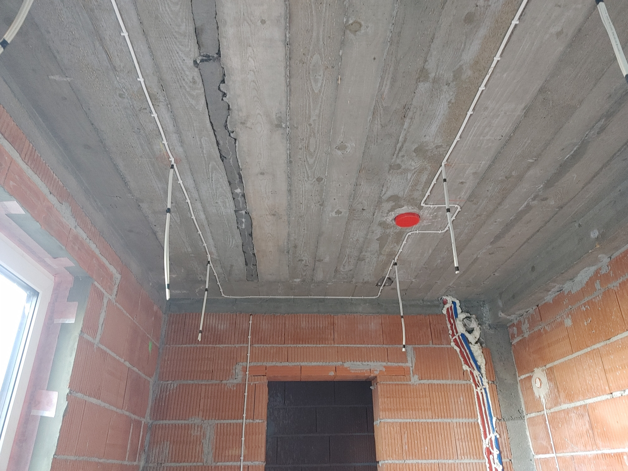 Surowy strop z widocznymi przewodami elektrycznymi prowadzonymi po powierzchni, mocowanymi uchwytami; ściany z czerwonej cegły dziurawki w trakcie budowy.
