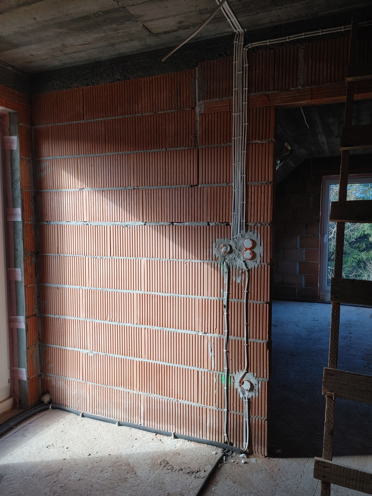 Instalacja elektryczna w surowym budynku: przewody w peszlach ułożone wzdłuż ściany z cegły kratówki, puszki podtynkowe zalane gipsem, widok na okno i drzewa w tle.