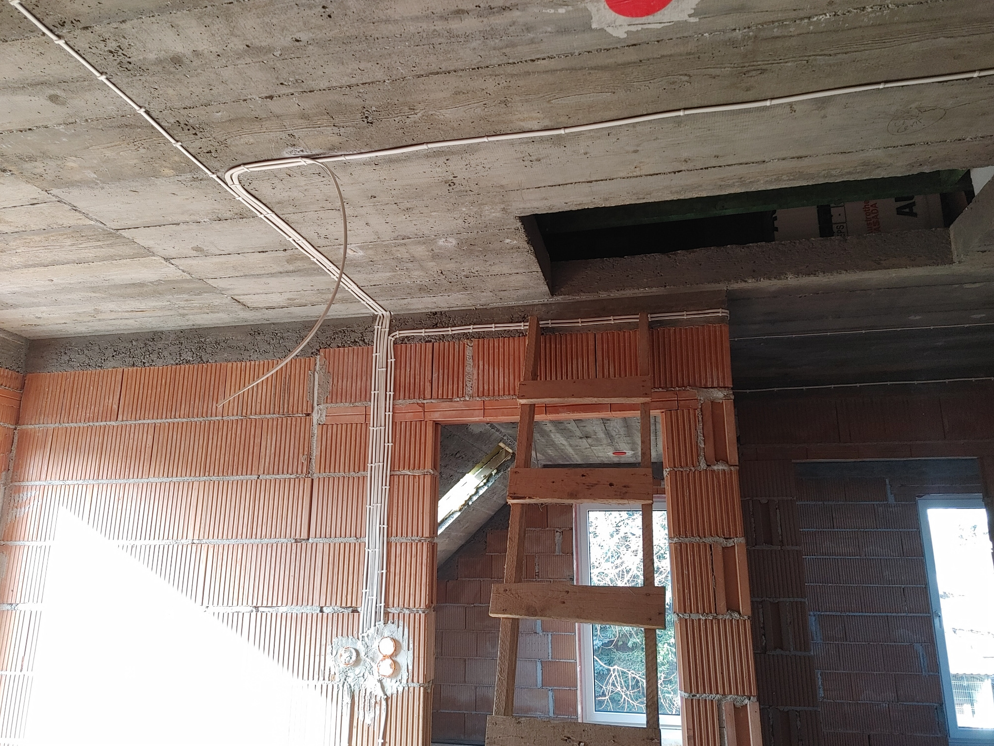 Surowy betonowy strop z widocznymi przewodami elektrycznymi w białych listwach, ściany z czerwonej cegły dziurawki w trakcie budowy, widoczna instalacja elektryczna w ścianach.