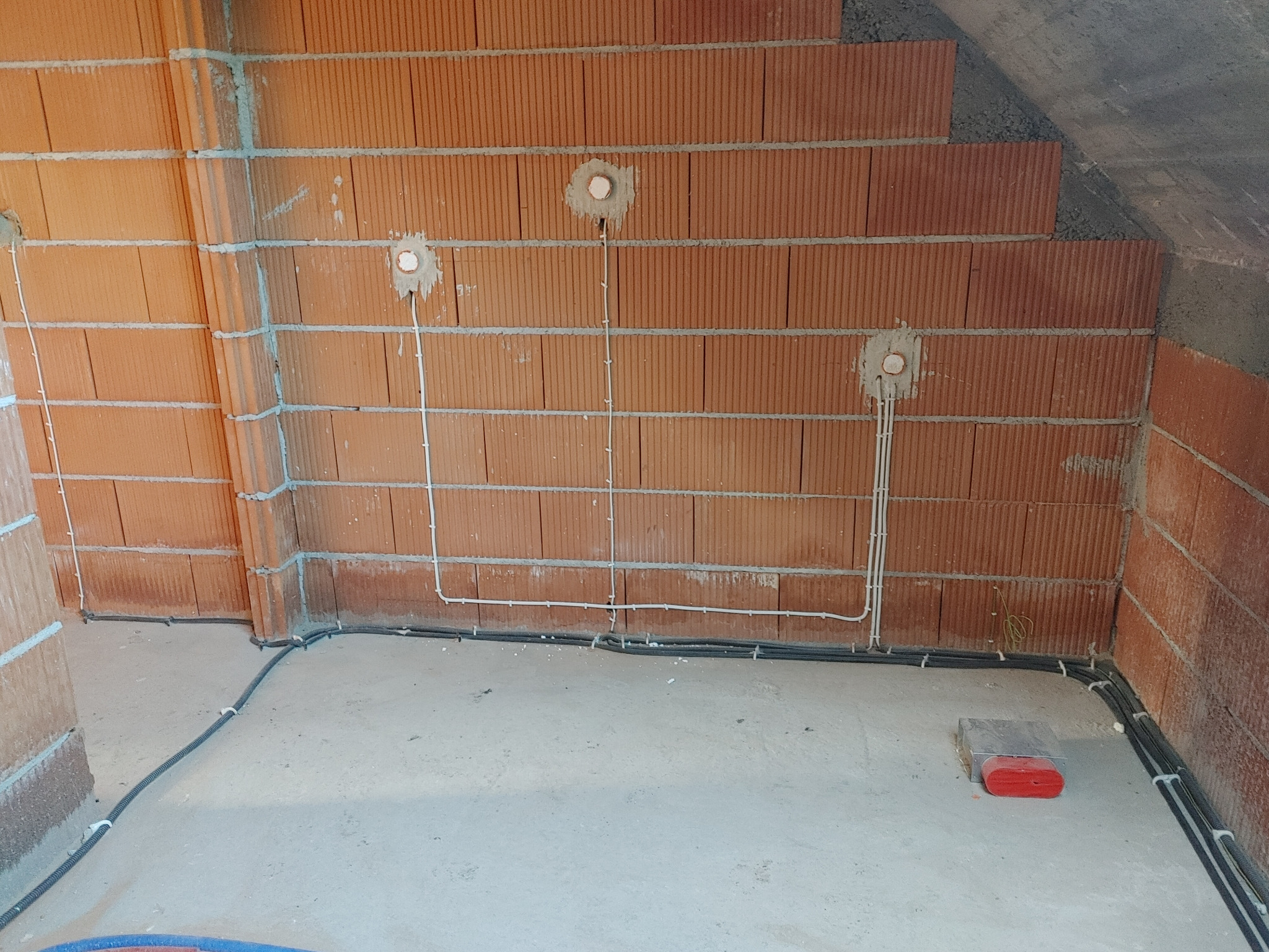 Wewnętrzne okablowanie elektryczne w surowym budynku z czerwonej cegły, widoczne przewody w ścianach i podłodze, przygotowane otwory pod gniazdka.