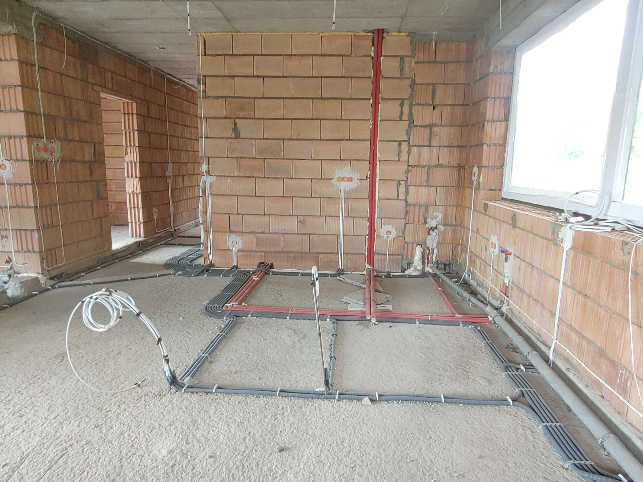 Surowy stan wnętrza budynku w trakcie rozprowadzania instalacji elektrycznej i rur, widoczne kable ułożone na podłodze i wyprowadzenia ze ścian z cegły.