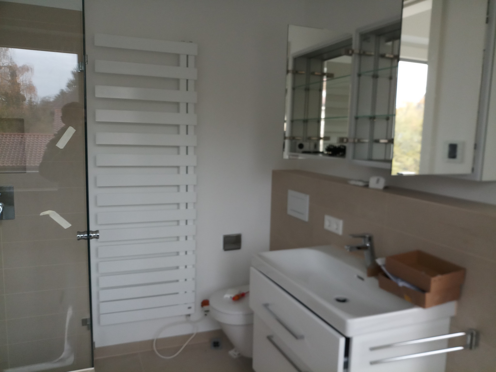 Nowoczesna łazienka z grzejnikiem drabinkowym, szafką z umywalką, lustrem z szafką i kabiną prysznicową, widoczne elementy instalacji elektrycznej - gniazdka.