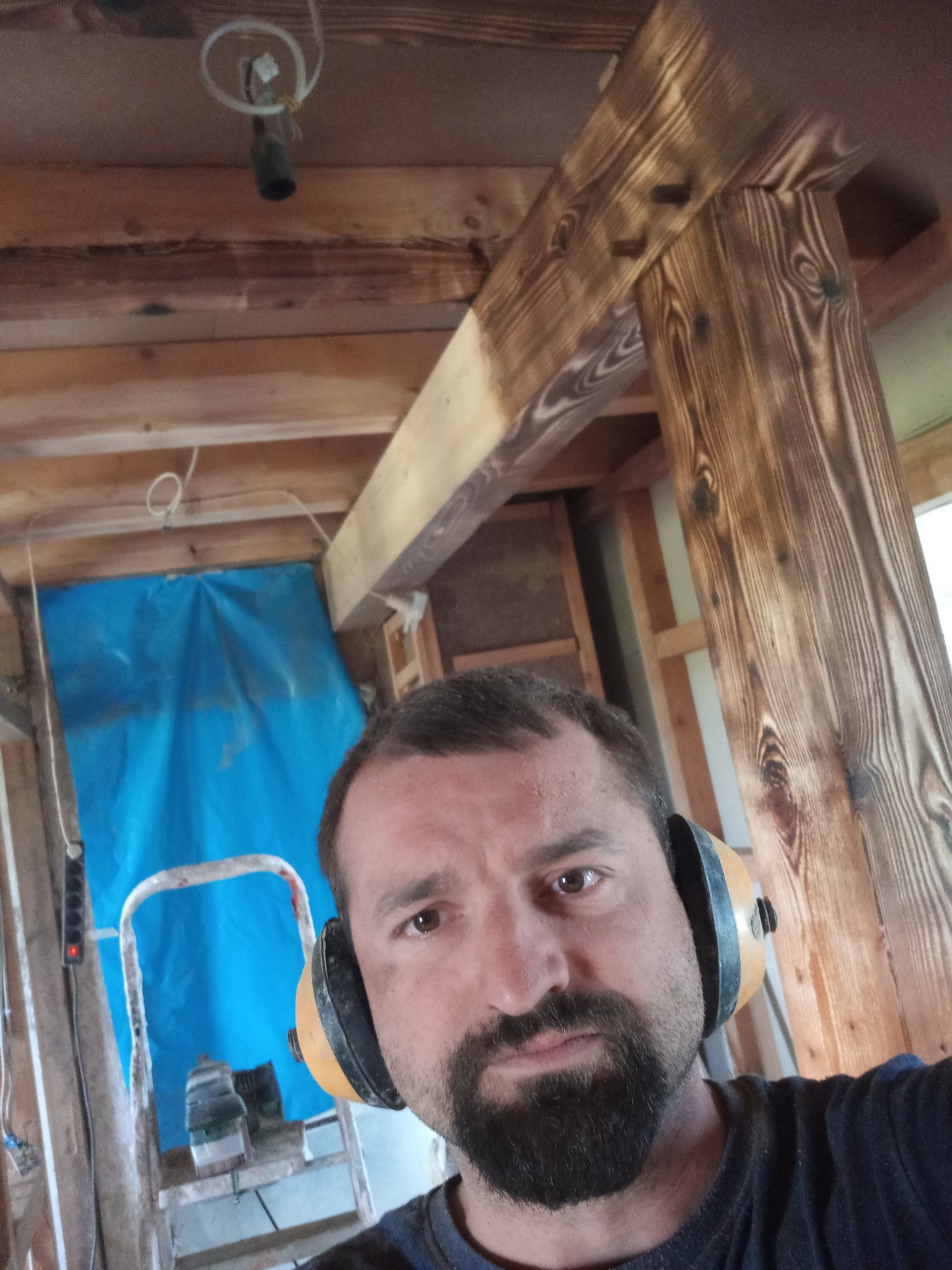 Mężczyzna w słuchawkach ochronnych, widoczne drewniane elementy konstrukcyjne budynku w trakcie budowy/remontu, częściowo opalane drewno, drabina i niebieska folia w tle.