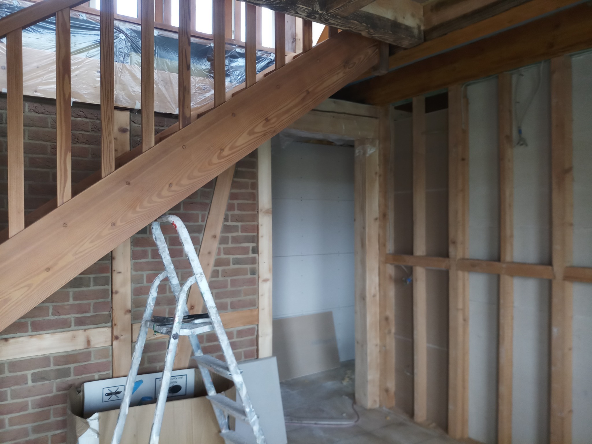 Wnętrze domu w trakcie remontu: drewniane schody z balustradą, ściany z cegły i drewnianych belek, aluminiowa drabina oparta o ścianę, widoczne przewody elektryczne na ścianie pokrytej płytami...