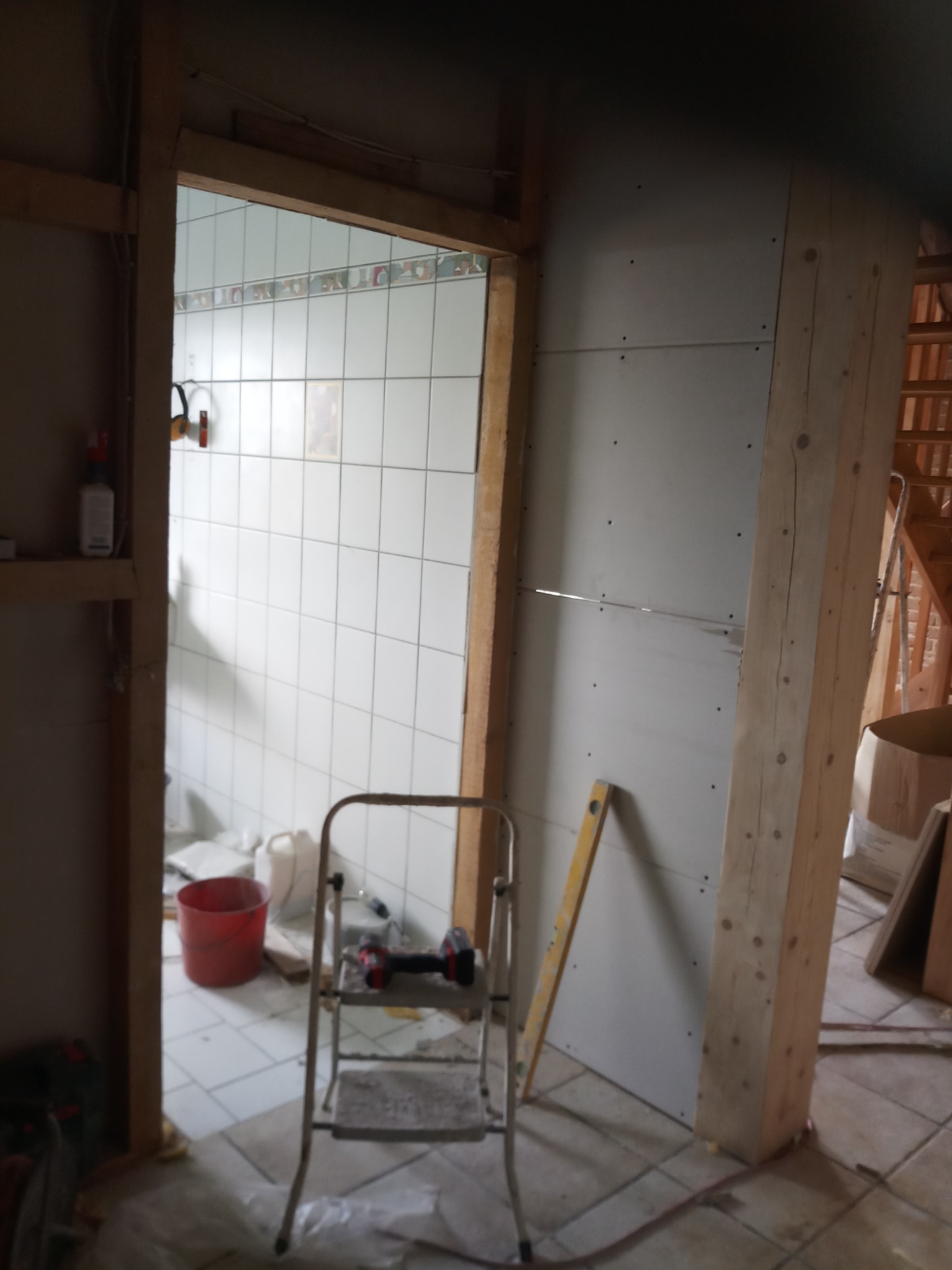 Wnętrze w trakcie remontu: widoczna ściana z płyt gipsowo-kartonowych, drewniana konstrukcja ramy drzwiowej, drabina aluminiowa z wiertarką akumulatorową na stopniu, poziomica i fragment...