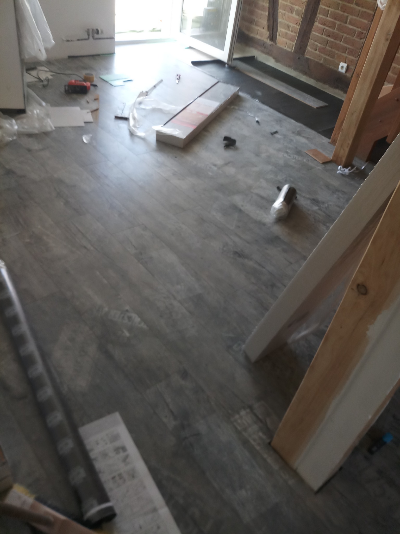 Montaż paneli podłogowych w pomieszczeniu z ceglaną ścianą, widoczna drabina malarska, narzędzia i materiały budowlane.