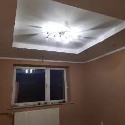 Wykończony sufit podwieszany z oświetleniem w pokoju o ścianach w kolorze terakoty, widoczny grzejnik pod oknem i materiały budowlane na podłodze.