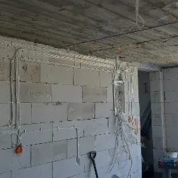 Wnętrze w stanie surowym z widoczną instalacją elektryczną w rurkach PCV, zamocowaną do ściany z bloczków betonowych i pod stropem, z rozdzielnicą elektryczną i puszkami pod gniazdka, z narzędziami...