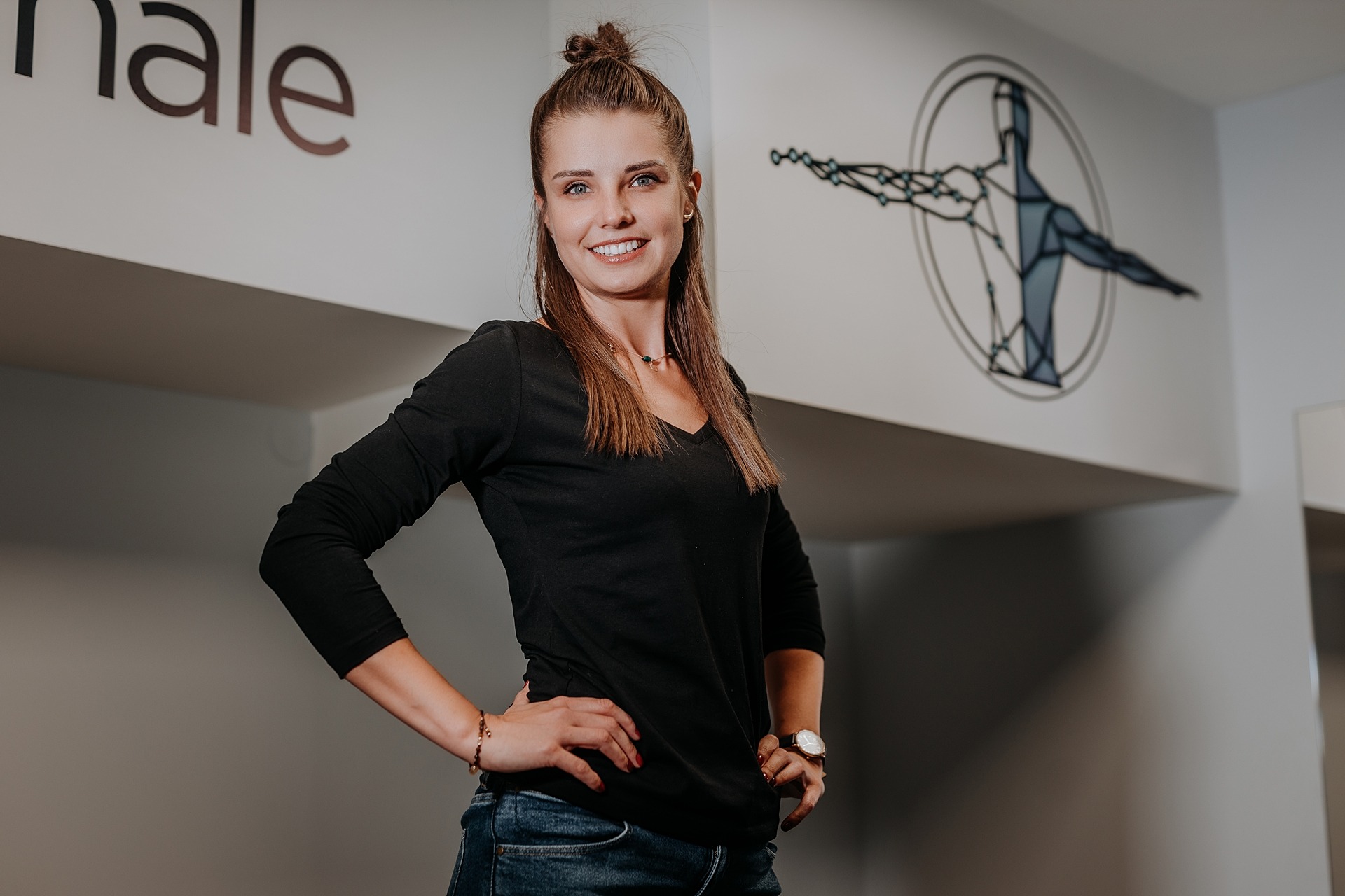 Uśmiechnięta kobieta w czarnej bluzce i jeansach stoi na tle białej ściany z dekoracyjnym rysunkiem sylwetki człowieka w okręgu.