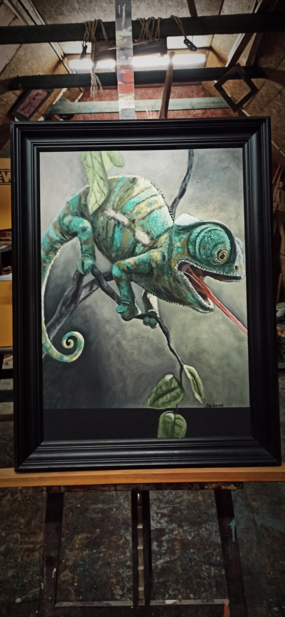 Oprawiony obraz przedstawiający kameleona z wyciągniętym językiem siedzącego na gałęzi, namalowany w odcieniach zieleni i błękitu na szarym tle.