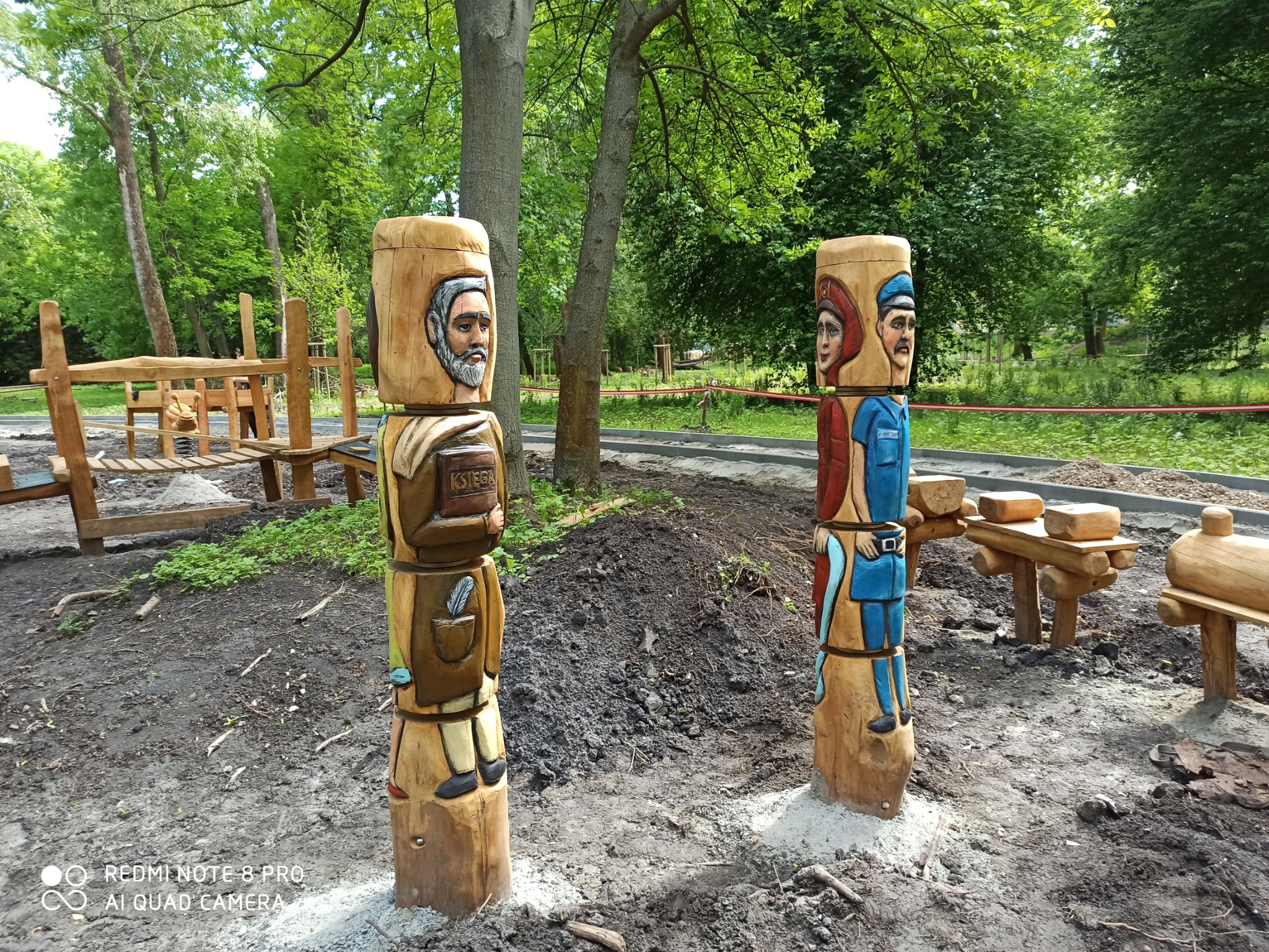 Ręcznie rzeźbione, drewniane figury postaci historycznych i bajkowych, umieszczone na placu zabaw z drewnianą konstrukcją w tle.