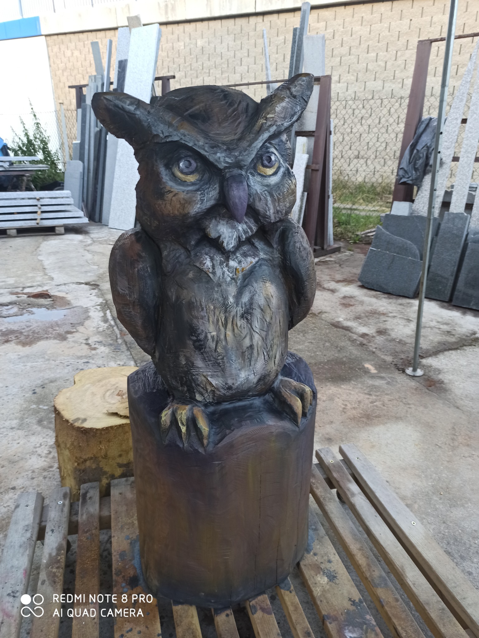 Drewniana rzeźba sowy z wyraźnymi śladami dłuta, postawiona na pniu w Nowym Targu, na tle płyt kamiennych i metalowego ogrodzenia.