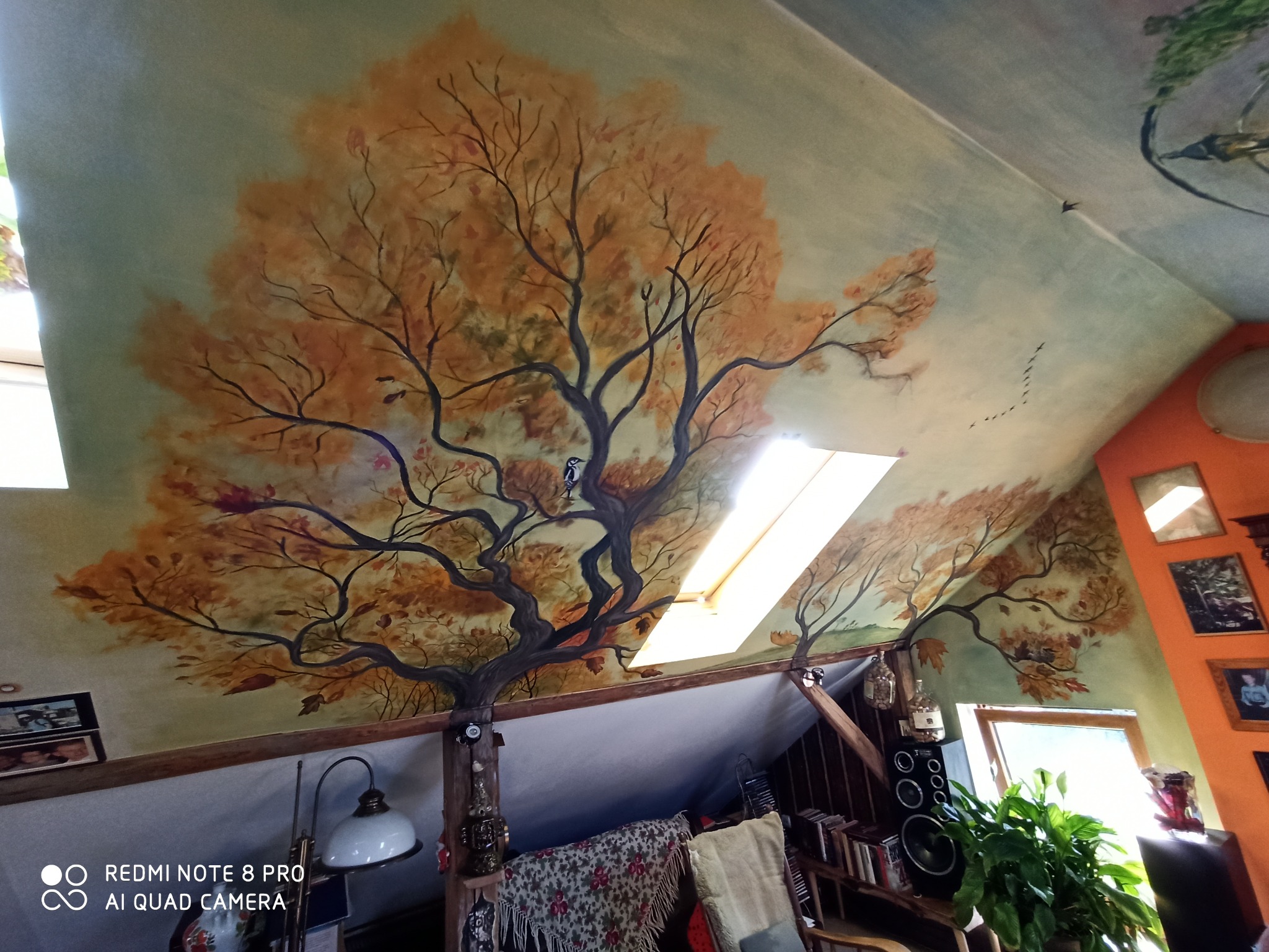 Malowidło na suficie poddasza przedstawiające drzewo w jesiennych barwach z ptakiem, z widocznym oknem dachowym i elementami wystroju wnętrza.