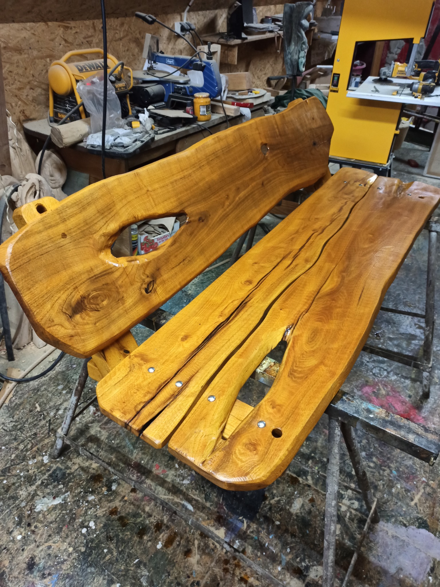 Dwie deski z jasnego drewna z widocznymi słojami i otworami, leżące na metalowym stojaku w warsztacie, z narzędziami i sprzętem w tle.