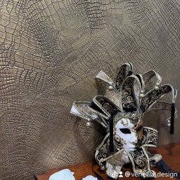 Venezia Design Stefano Callegaro - Dekoracyjna ściana z fakturą skóry krokodyla w odcieniach złota, obok leży wenecka maska karnawałowa w kolorach czerni, bieli i złota, z dzwoneczkami.
