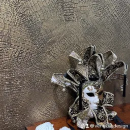 Dekoracyjna ściana z fakturą skóry krokodyla w odcieniach złota, obok leży wenecka maska karnawałowa w kolorach czerni, bieli i złota, z dzwoneczkami.