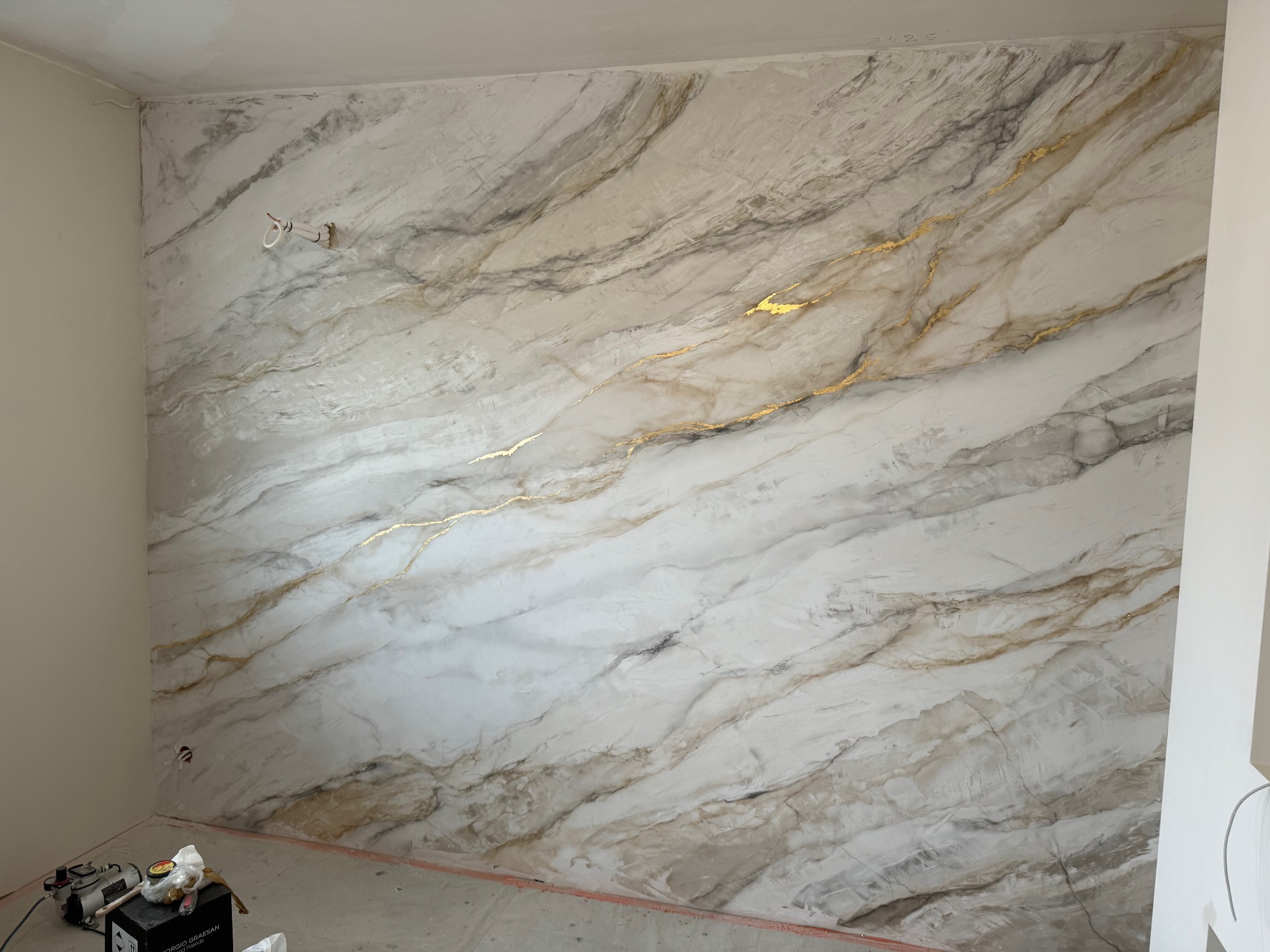 Ściana wewnętrzna z dekoracyjnym tynkiem imitującym marmur z żyłkami w odcieniach złota, widoczne niedokończone prace wykończeniowe.
