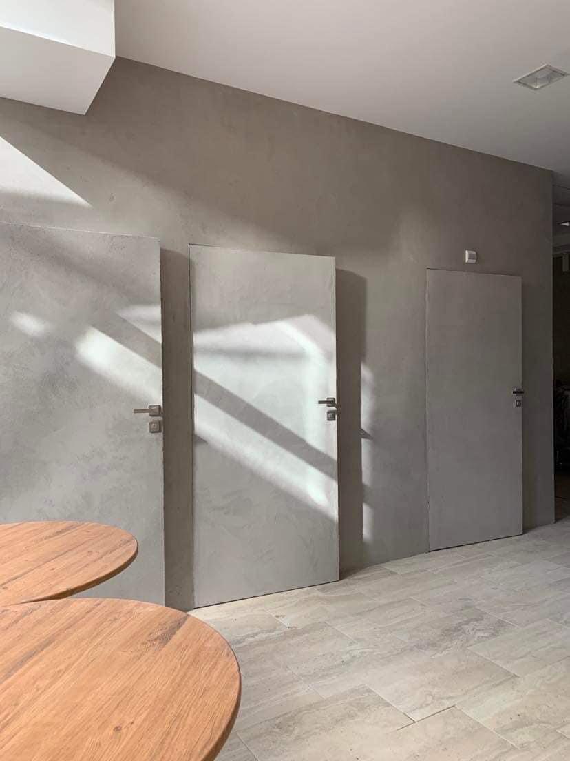 Wnętrze z trzema ukrytymi drzwiami w ścianie o fakturze betonu, oświetlonymi naturalnym światłem, z fragmentami drewnianych okrągłych stolików w kadrze.