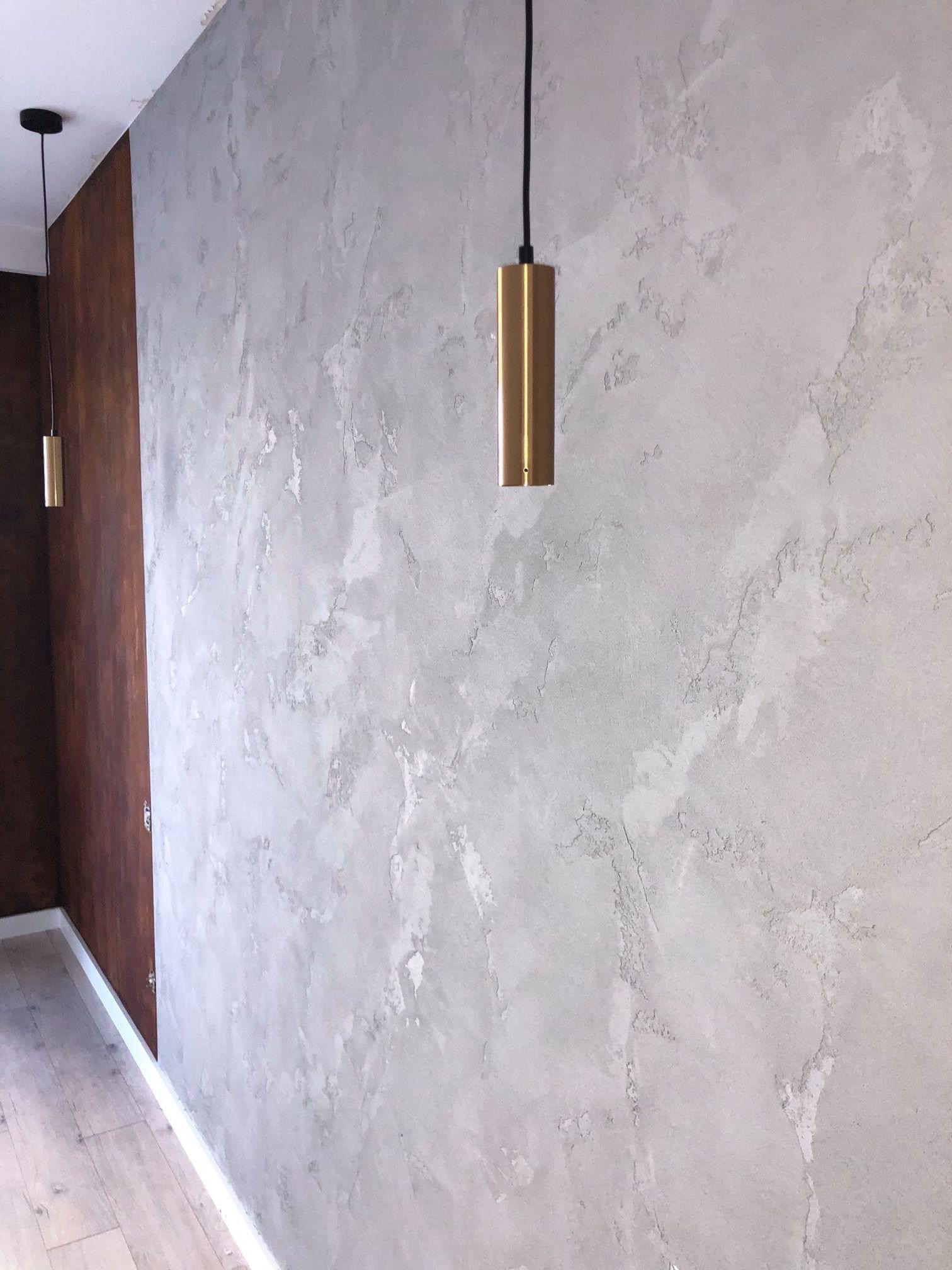 Szara ściana z efektem dekoracyjnym, oświetlona złotą lampą wiszącą, sąsiadująca z ciemnobrązową okładziną ścienną.