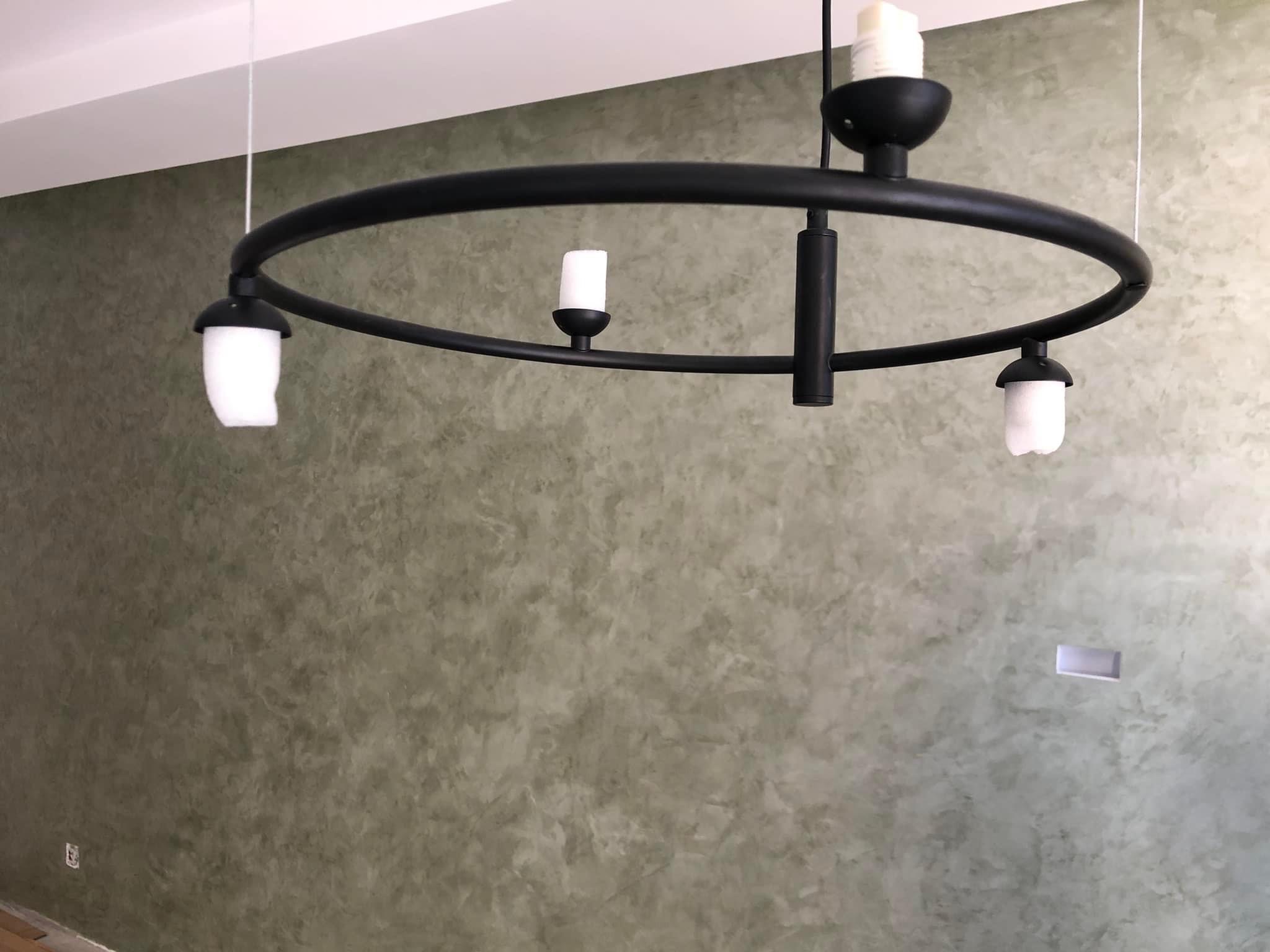 Ściana z dekoracyjną strukturą w odcieniach zieleni i szarości, oświetlona przez nowoczesną, okrągłą lampę wiszącą z czarnym metalowym stelażem i trzema białymi kloszami.