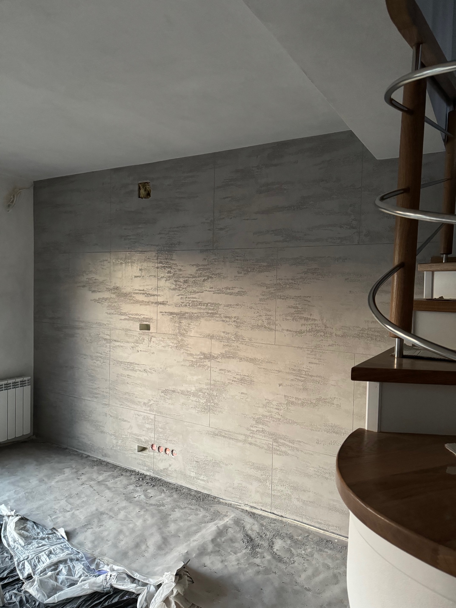 Ściana wykończona dekoracyjnym tynkiem w odcieniach szarości w Jędrzejowie, widoczne gniazdka elektryczne i fragment schodów spiralnych z drewnianymi stopniami, podłoga zabezpieczona folią malarską.