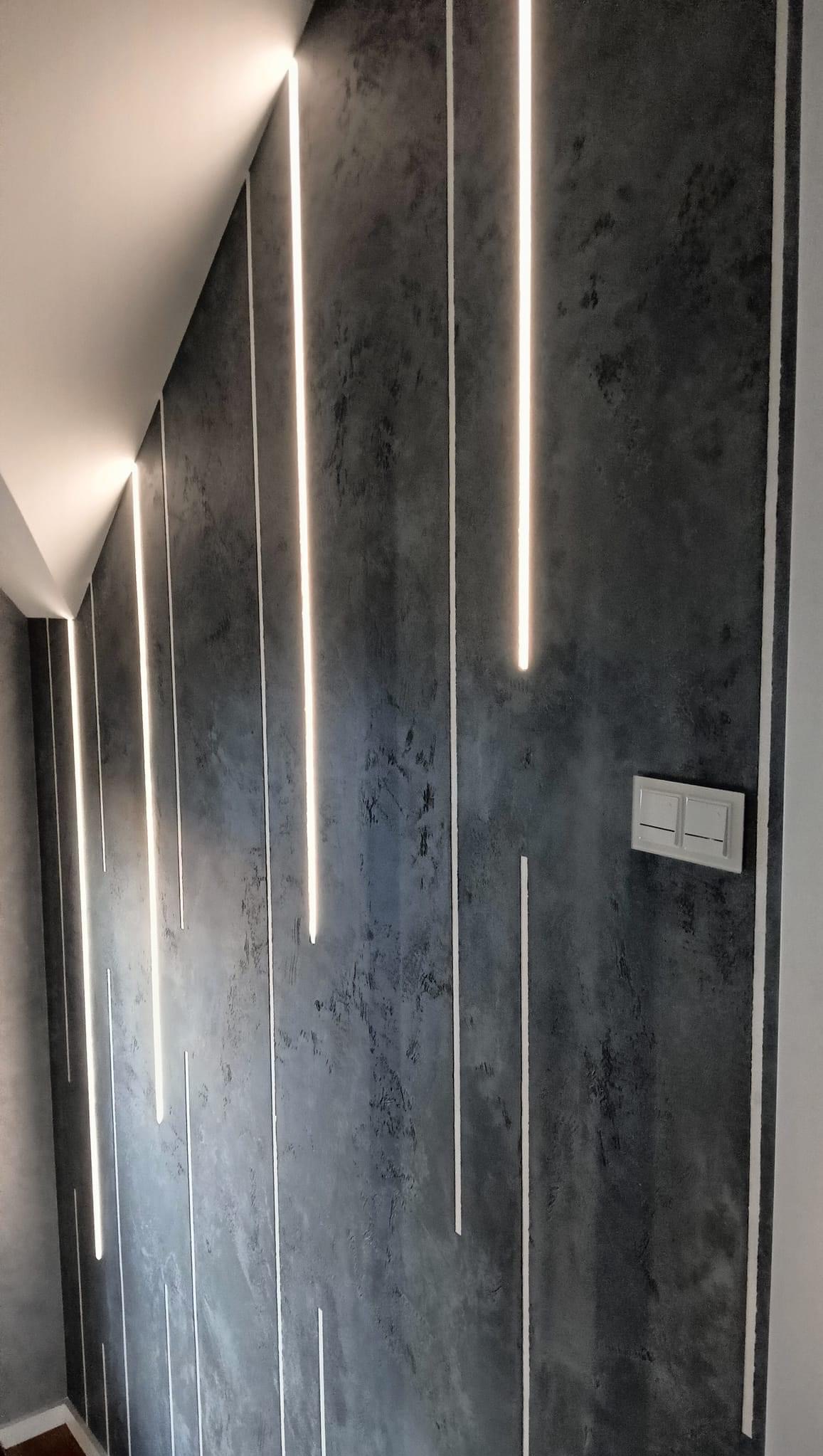 Szara ściana z dekoracyjnym tynkiem i pionowymi paskami LED, widoczny włącznik światła.