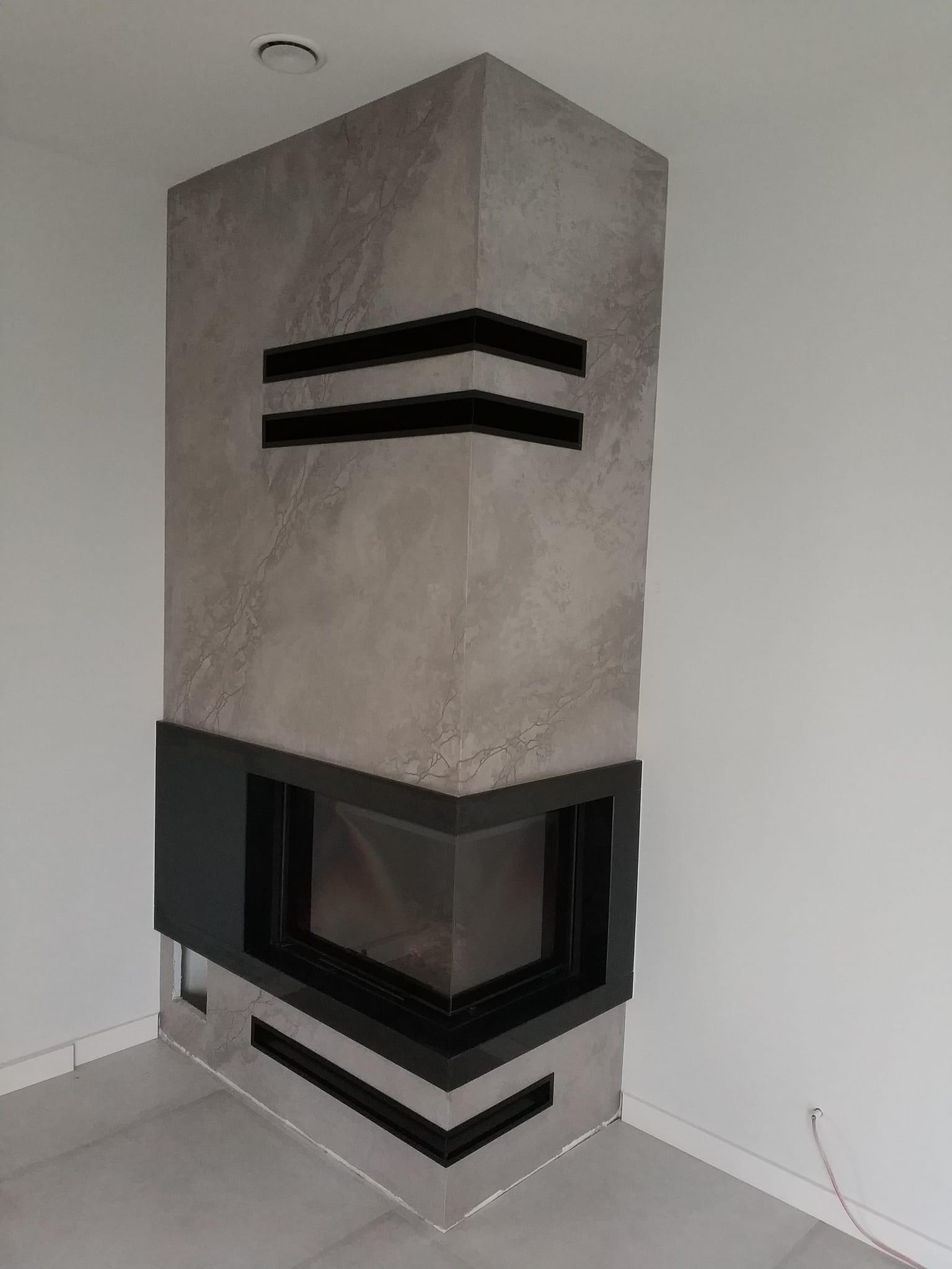 Nowoczesny kominek narożny z obudową w odcieniach szarości z czarnymi akcentami, w minimalistycznym wnętrzu.