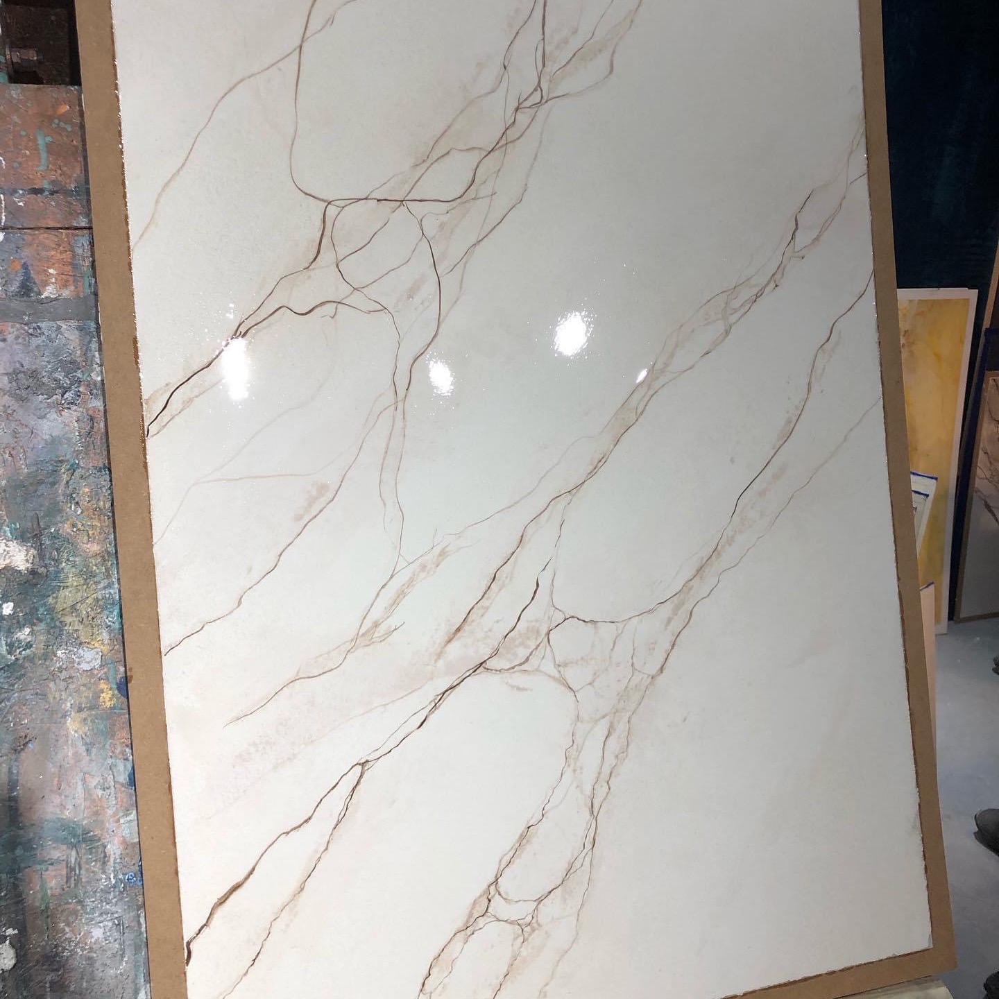 Panel z imitacją marmuru w odcieniach bieli i beżu, oparty o malarską sztalugę, widoczne refleksy światła na powierzchni.