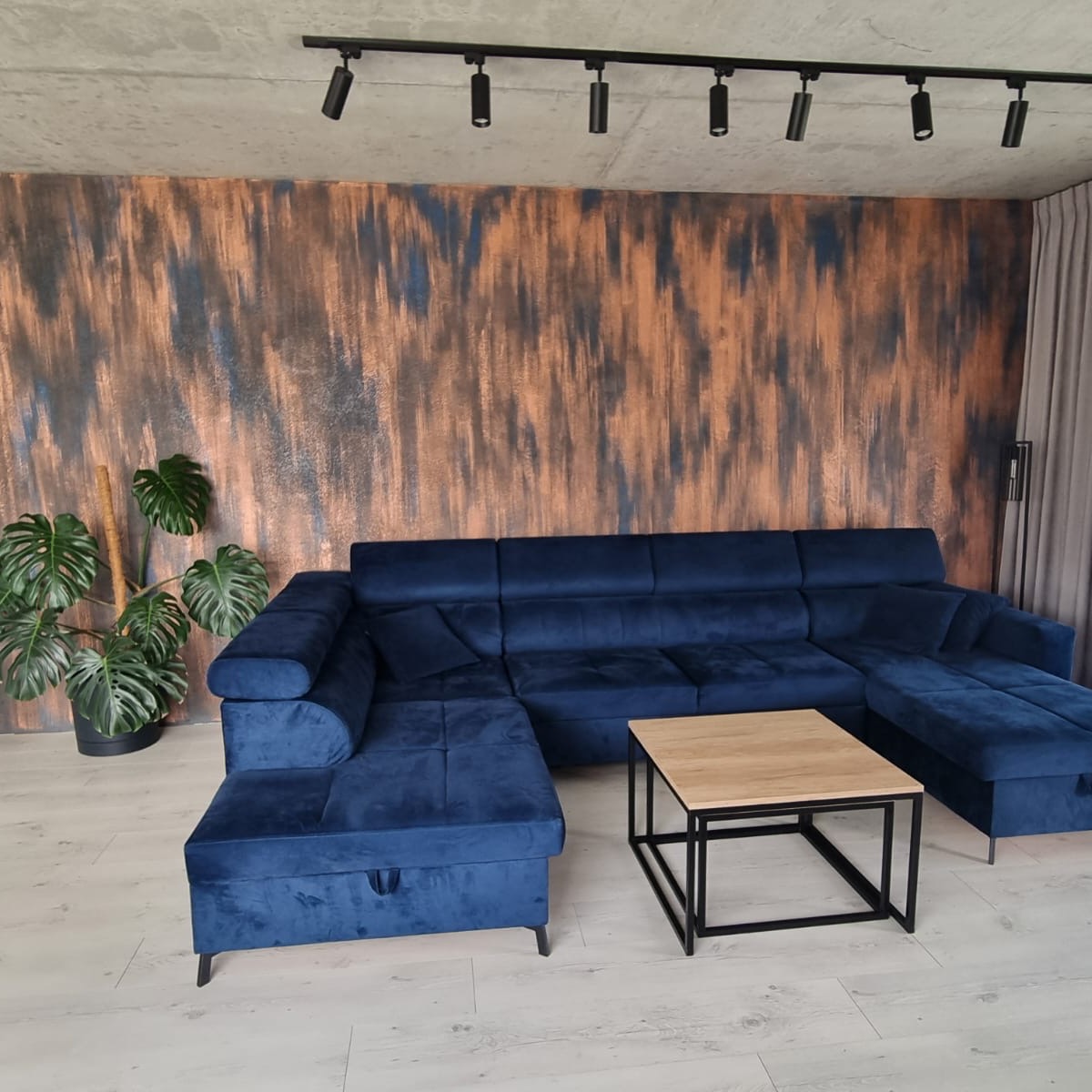 Granatowy, welurowy narożnik z pufą i stolikiem kawowym na tle ściany z efektem rdzy, oświetlony reflektorami.