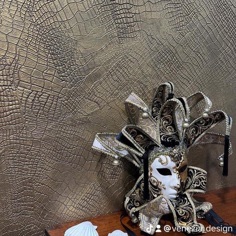 Dekoracyjna ściana z fakturą skóry krokodyla w odcieniach złota, obok leży wenecka maska karnawałowa w kolorach czerni, bieli i złota, z dzwoneczkami.