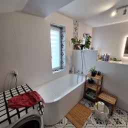 Jasna łazienka z wolnostojącą wanną, biało-czarną mozaiką na podłodze i oknem z żaluzjami. Pralka z narzuconą tkaniną w kratę stoi obok wanny.