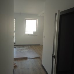 LENBUD Ogólnobudowlane - Wnętrze pokoju z drewnianą podłogą, częściowo pokrytą folią ochronną z wzorem liści palmowych, widok z korytarza przez otwarte drzwi.