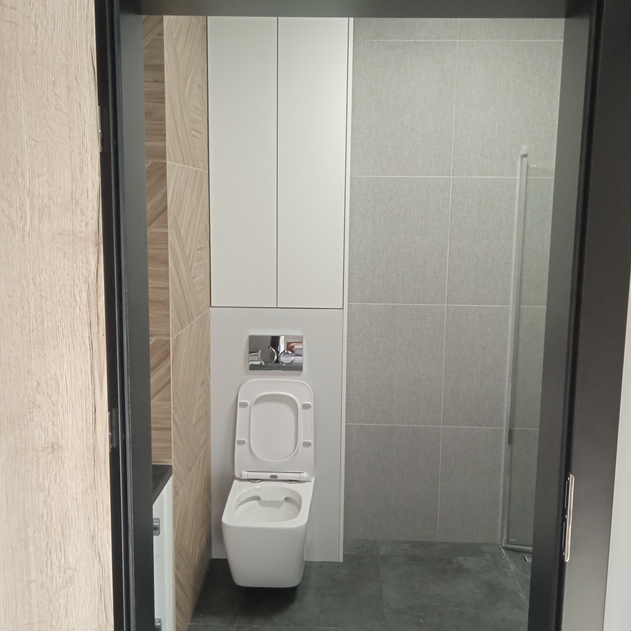 Nowoczesna łazienka z podwieszaną toaletą, zabudowaną szafką nad stelażem, szarą glazurą i kabiną prysznicową typu walk-in, widziana zza ciemnej futryny drzwi.