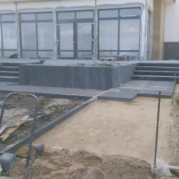 Budowa tarasu z kamiennymi schodami i przygotowanym podłożem z piasku. Widoczna zagęszczarka i narzędzia budowlane.