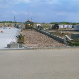 Prace brukarskie w toku: przygotowanie podłoża, ustawianie obrzeży, wyrównywanie terenu za pomocą niwelatora i miniładowarki. W tle budynek w stanie surowym otwartym oraz materiały budowlane.