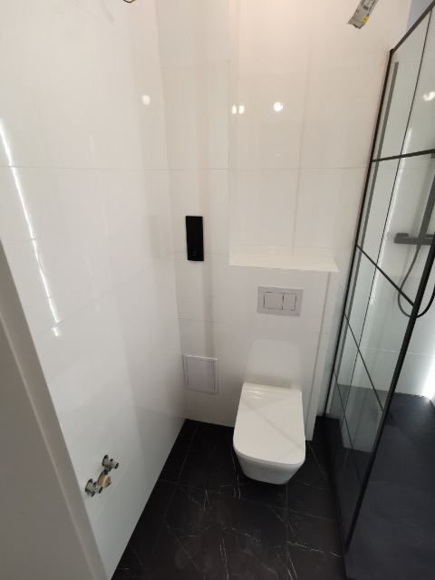 Nowoczesna łazienka z białymi płytkami, czarną podłogą imitującą marmur i minimalistyczną toaletą podwieszaną.