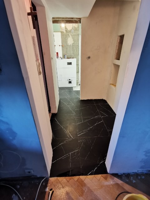 Wykończona podłoga w łazience wyłożona czarnymi płytkami z białym żyłkowaniem, widok z korytarza na odnowione wnętrze, ściany przygotowane do malowania, w tle biały bojler.