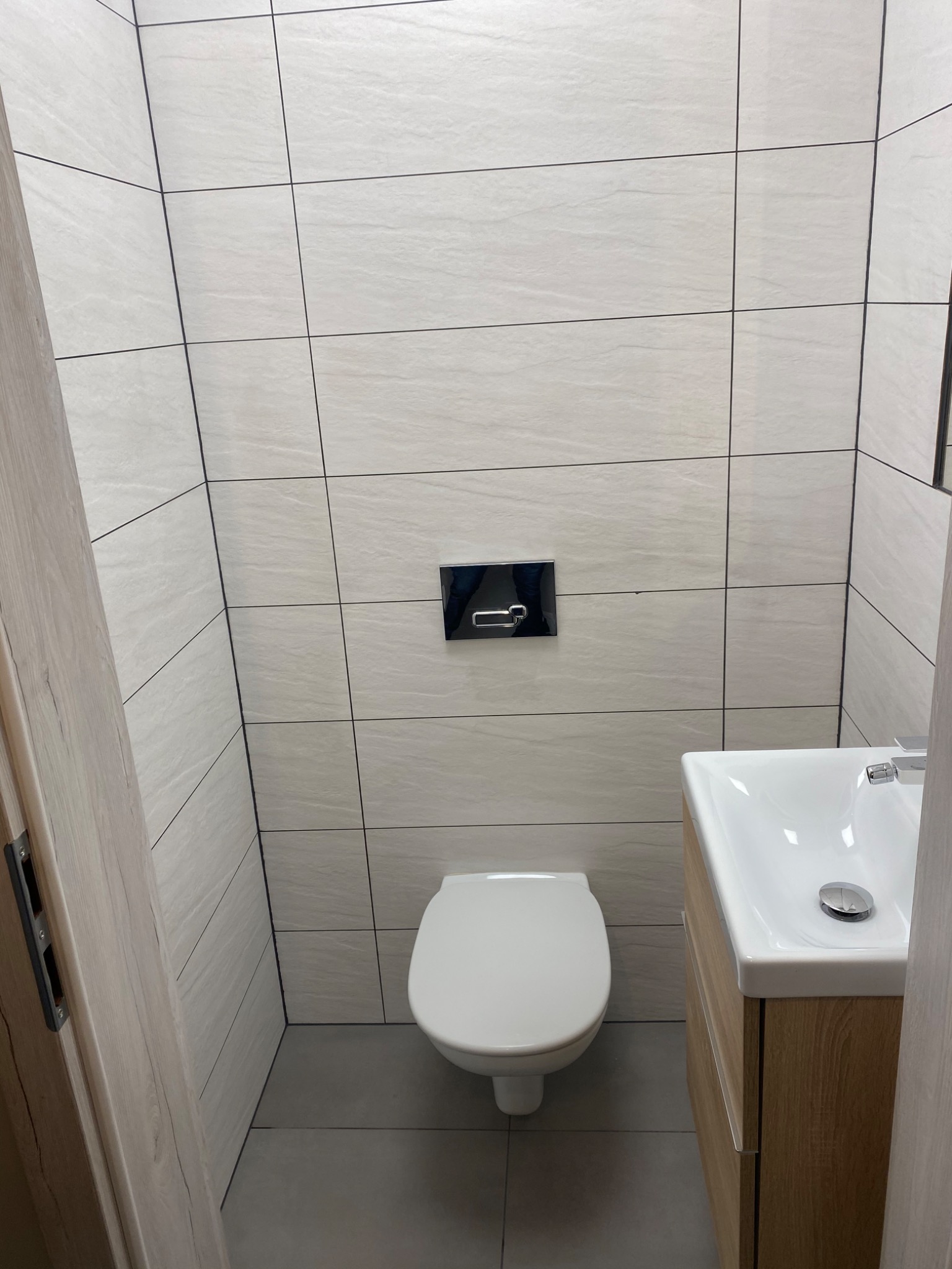 Wykończona łazienka z beżowymi płytkami imitującymi kamień, podwieszaną toaletą i umywalką w szafce z drewnopodobnym fornirem, widok zza uchylonych drzwi.