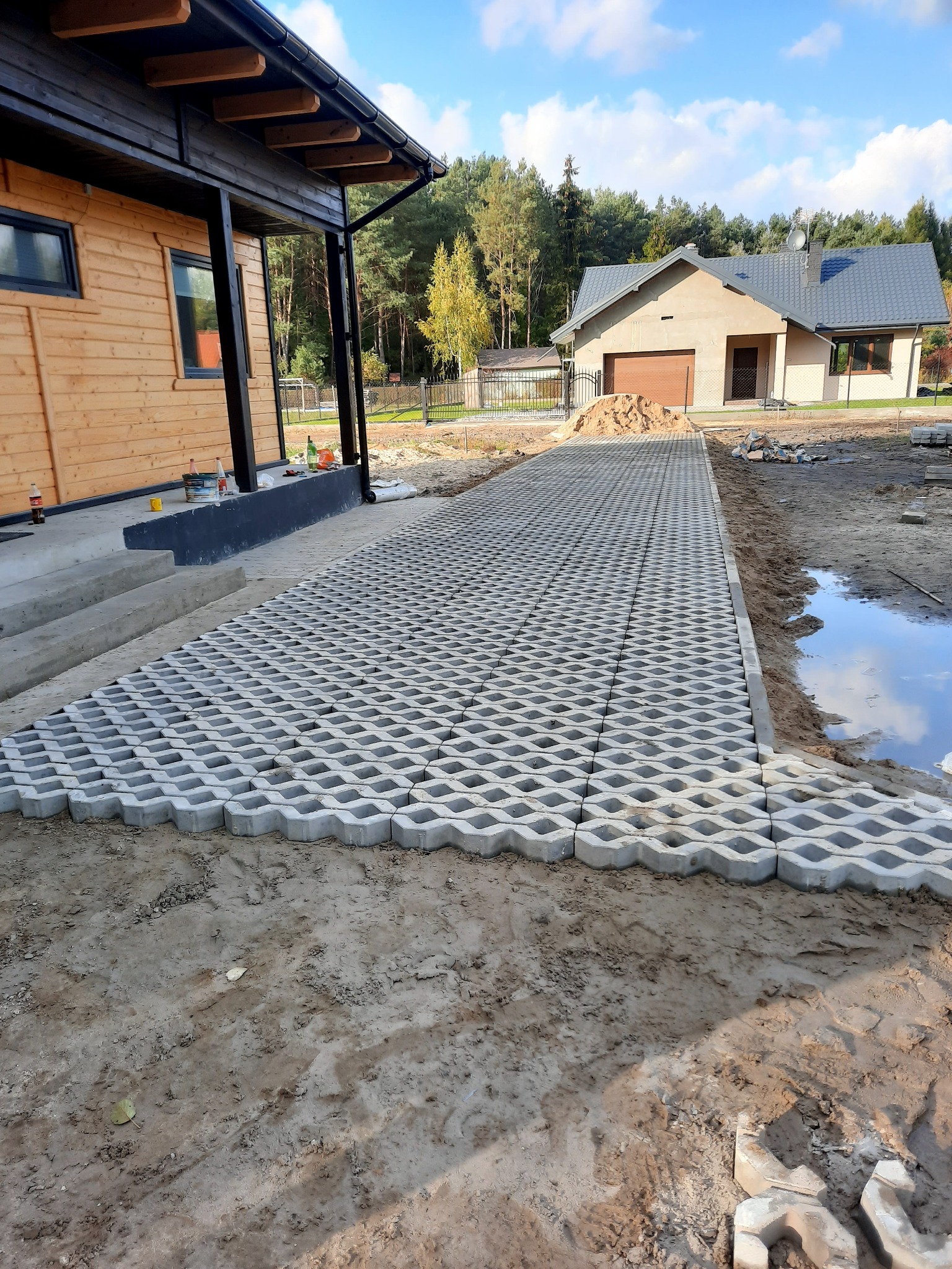 Układanie przepuszczalnej kostki brukowej typu plaster miodu na podjeździe, z widocznym budynkiem mieszkalnym w tle i drewnianym domkiem z boku, częściowo ułożona nawierzchnia na piaszczystym podłożu.
