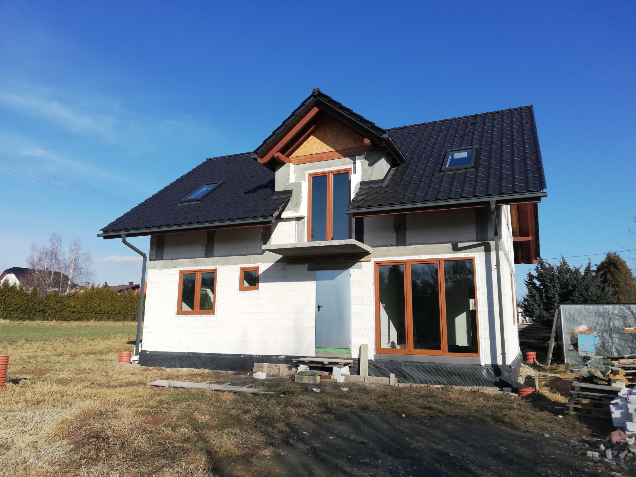 Niedokończony dom jednorodzinny z białych bloczków, czarnym dachem z oknami dachowymi i brązowymi ramami okiennymi w słoneczny dzień. Widoczne materiały budowlane i tymczasowe wejście.