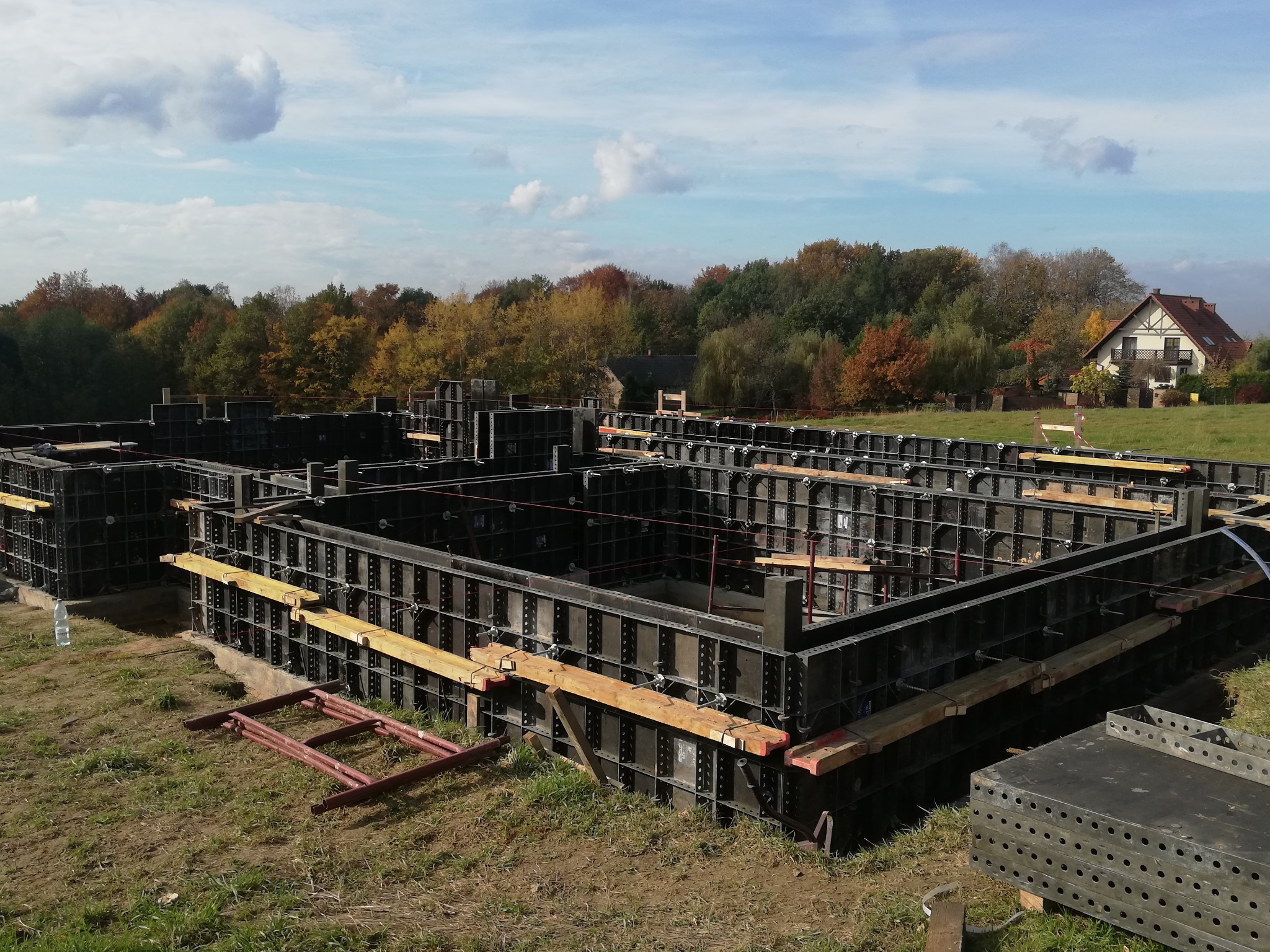 Szare szalunki fundamentowe z drewnianymi wzmocnieniami na trawiastym terenie, w tle jesienny las i dom jednorodzinny pod błękitnym niebem z chmurami.