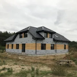 Nowoczesny dom dwukondygnacyjny z szarą elewacją, poziomym pasem z drewnianych paneli i czarnym dachem, otoczony naturalnym krajobrazem z lasem w tle.