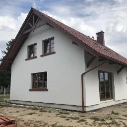 Świeżo otynkowany dom jednorodzinny z brązową dach&oacute;wką i drewnianymi elementami wykończeniowymi, widoczny z boku w pochmurny dzień.