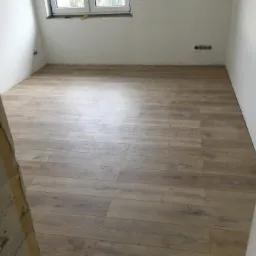 Nowo ułożone panele podłogowe w jasnym odcieniu drewna w pustym pokoju z białymi ścianami, widoczne gniazdka elektryczne i surowy fragment ściany przy wejściu.