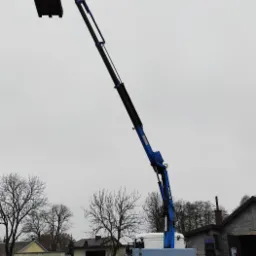 Niebieski dźwig samochodowy MAN podnosi paletę z ładunkiem na tle szarego nieba i zabudowań gospodarczych. Wokół leżą stosy drewnianych palet.