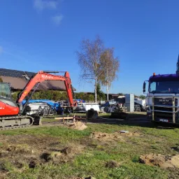 Czerwona mini koparka Kubota KX080-4 i biała betoniarka MAN na placu budowy w słoneczny dzień, w tle dom z panelami słonecznymi.