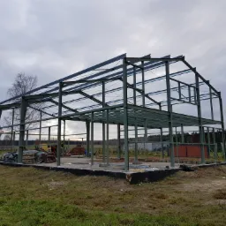 Konstrukcja stalowa hali w trakcie montażu, widoczne słupy i belki, przygotowany fundament, teren budowy z zieloną trawą i drzewami w tle, pochmurne niebo.