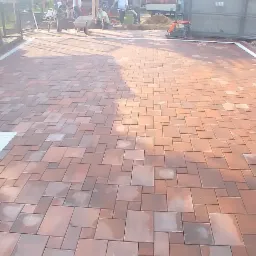 Nowo ułożona, brązowo-czerwona kostka brukowa o nieregularnym kształcie, z białym obramowaniem, częściowo mokra, z widocznymi pracownikami w tle i ubijarką wibracyjną.